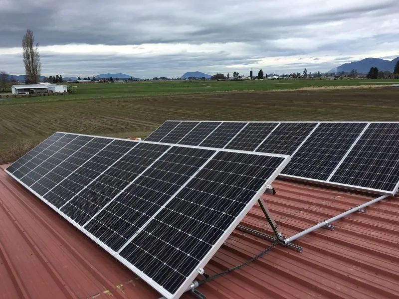 Solar panels installed on a red metal roof overlooking farmland and distant mountains under a cloudy sky.