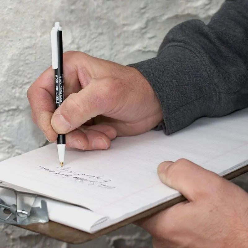 Person writing on clipboard with pen, against a stone wall background.