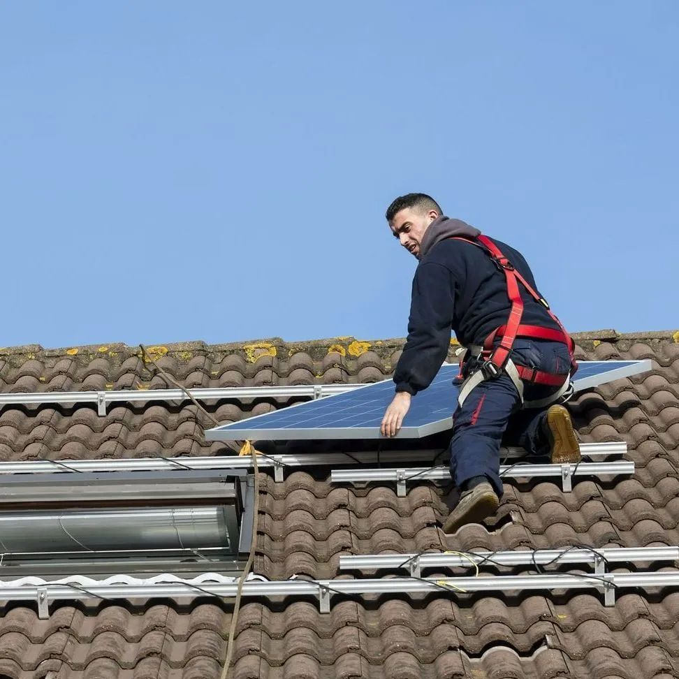 Roofer in safety harness installing solar panel on a brown-tiled roof under a blue sky.