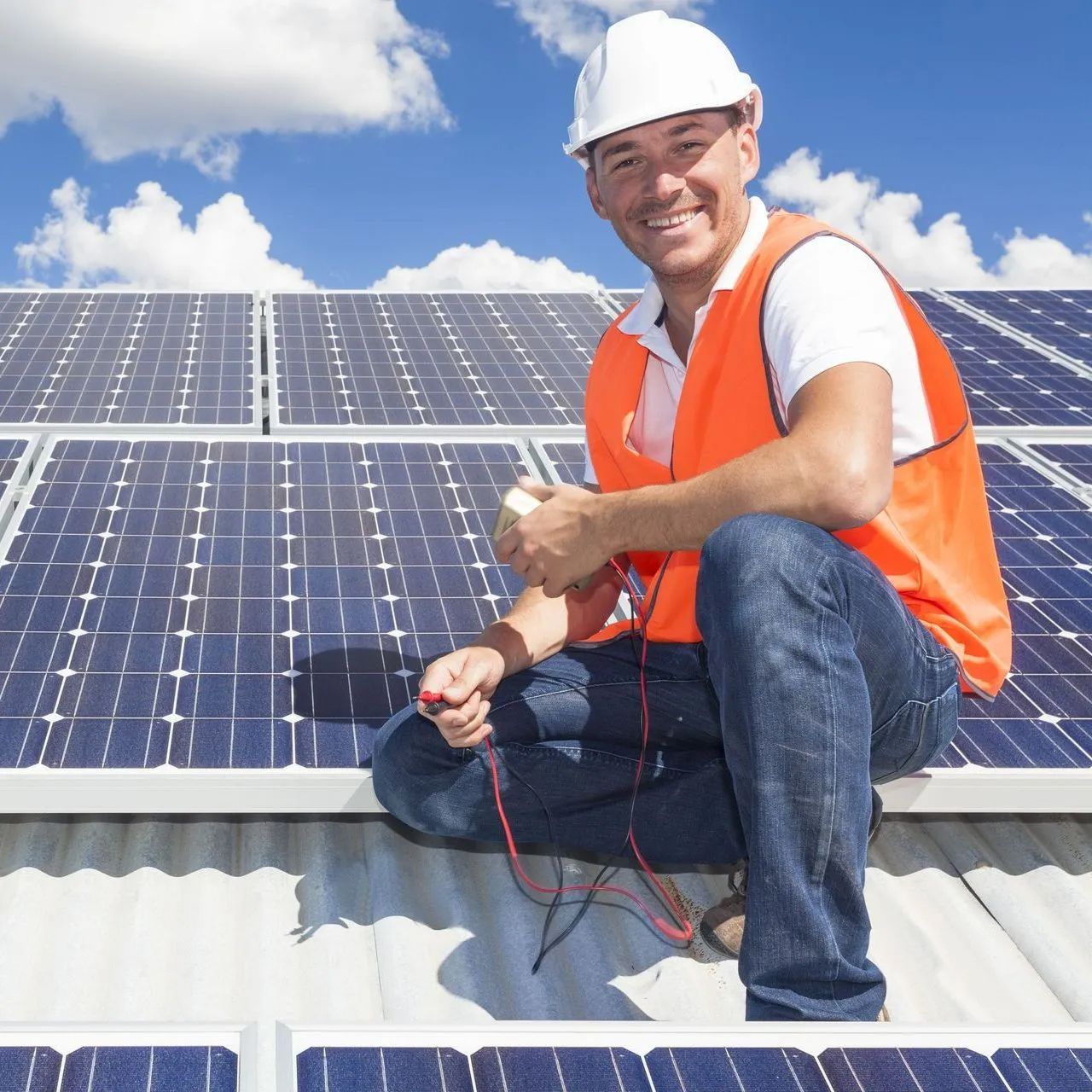 Solar panel technician in orange vest and hard hat, smiling, kneeling on rooftop, checking equipment.
