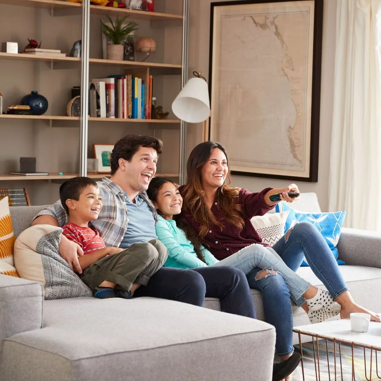 Family watches TV on a couch, living room. Parents and two children smile. One holds the remote.