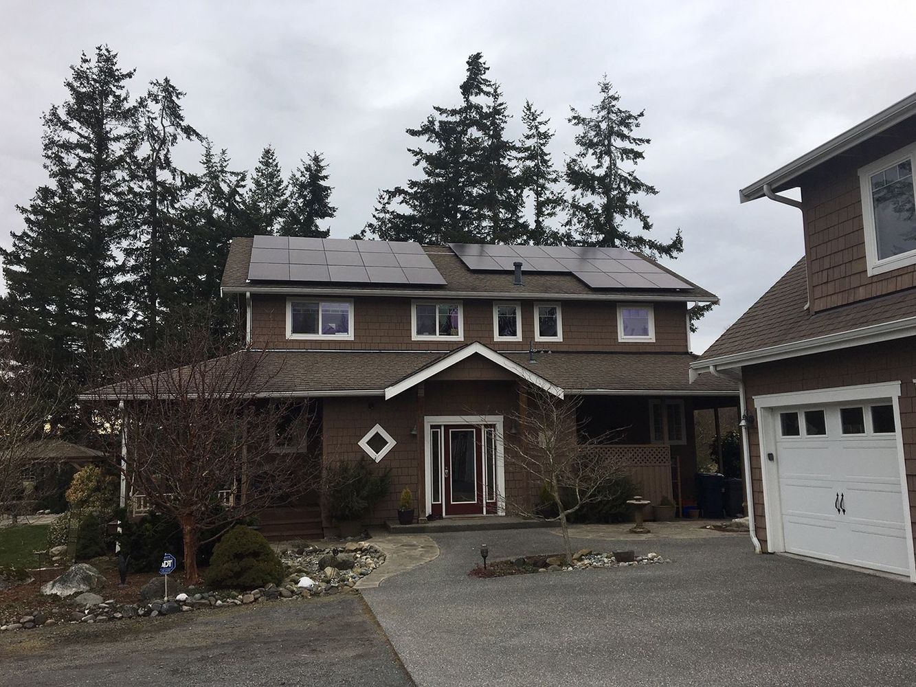 Brown house with solar panels on roof, under cloudy sky. Driveway, trees in the background.