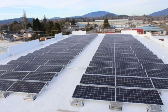 Solar panels arranged in rows on a white rooftop. Mountains and buildings in the background under a blue sky.