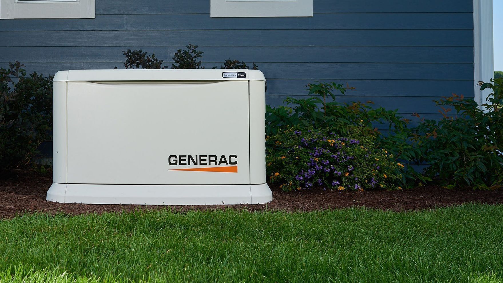 A Generac home standby generator on a grassy lawn next to a blue house with bushes.