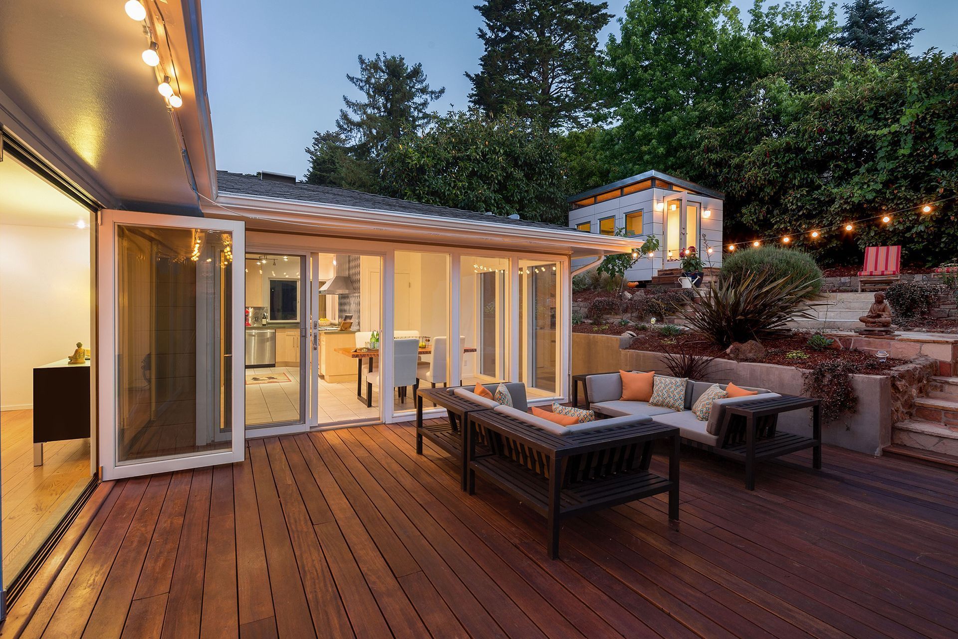 Wooden deck with patio furniture, open doors to a kitchen, string lights, and a garden with trees.