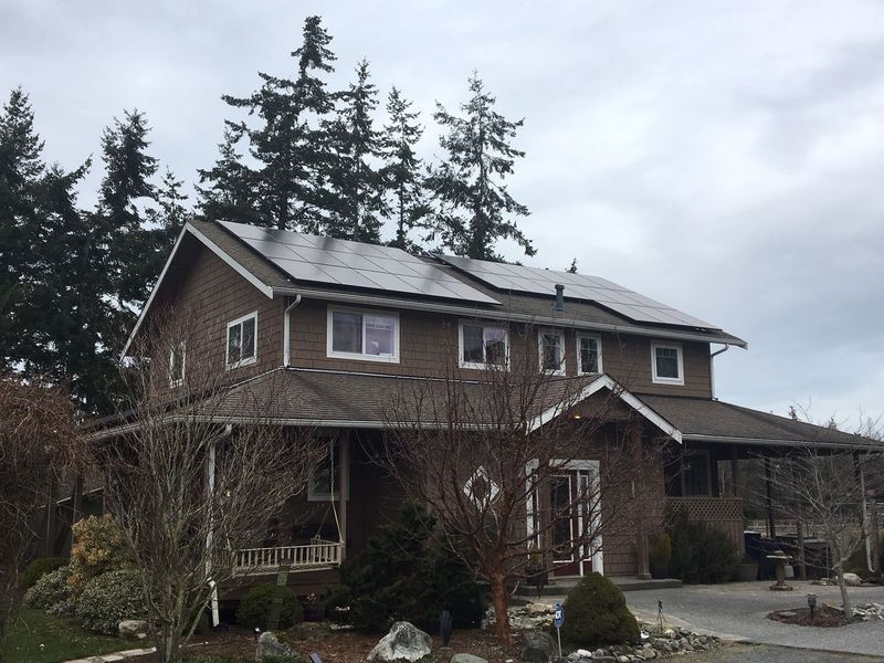 Brown two-story house with solar panels on roof under a cloudy sky, surrounded by trees and bushes.