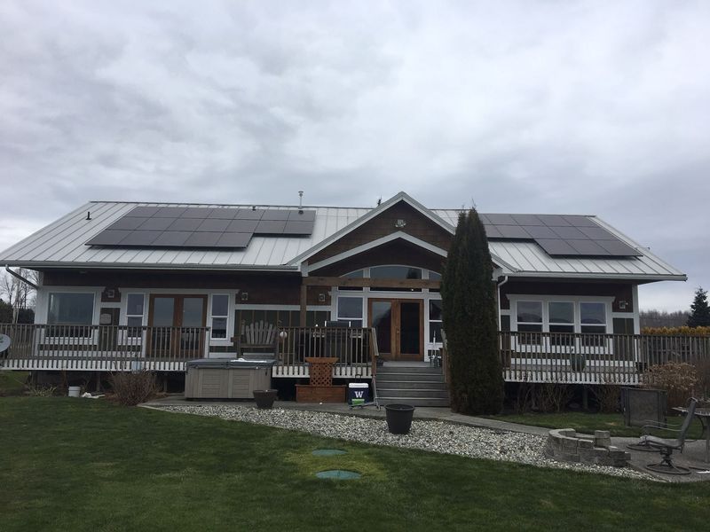 House with solar panels on a silver metal roof, brown siding, and deck. Overcast sky.