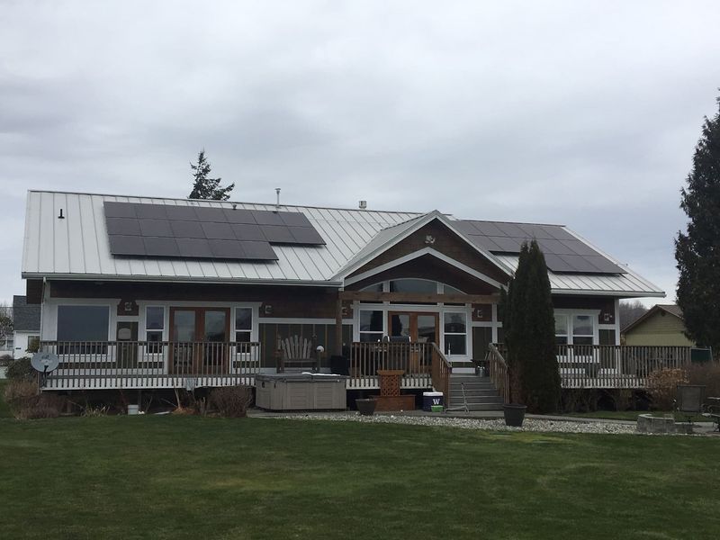 House with solar panels on a silver metal roof, brown siding, and a wrap-around deck.