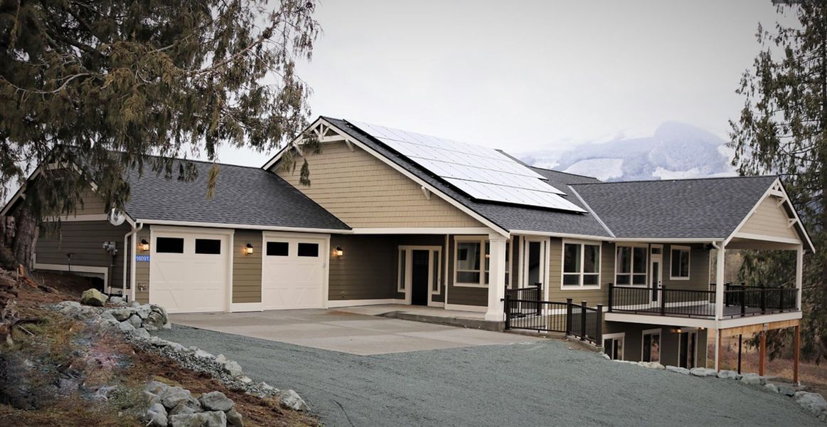 House with solar panels on roof, two-car garage, and mountain backdrop on a cloudy day.