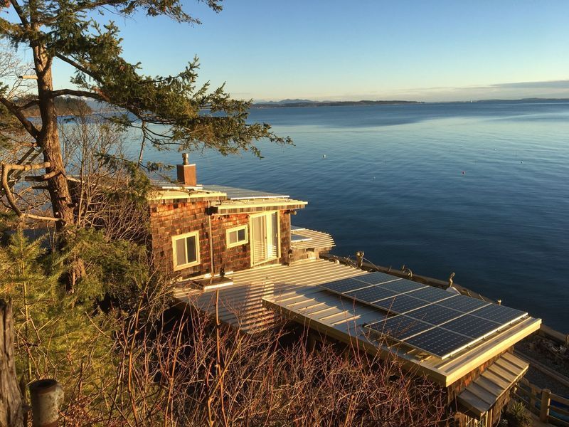 Cottage with solar panels on a bluff overlooking a calm, blue ocean. Sunny day, with trees nearby.