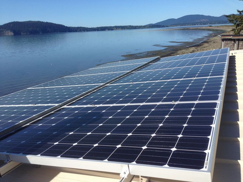 Solar panels on a roof overlooking water and a distant shoreline under a bright blue sky.