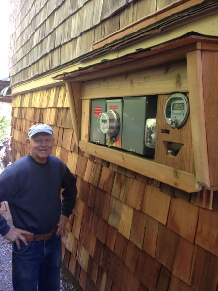 Man stands beside an outdoor electrical meter box mounted on a cedar shingle wall.