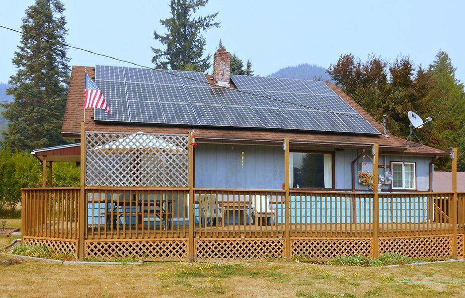 Blue house with solar panels, wooden deck, American flag, and mountain backdrop.