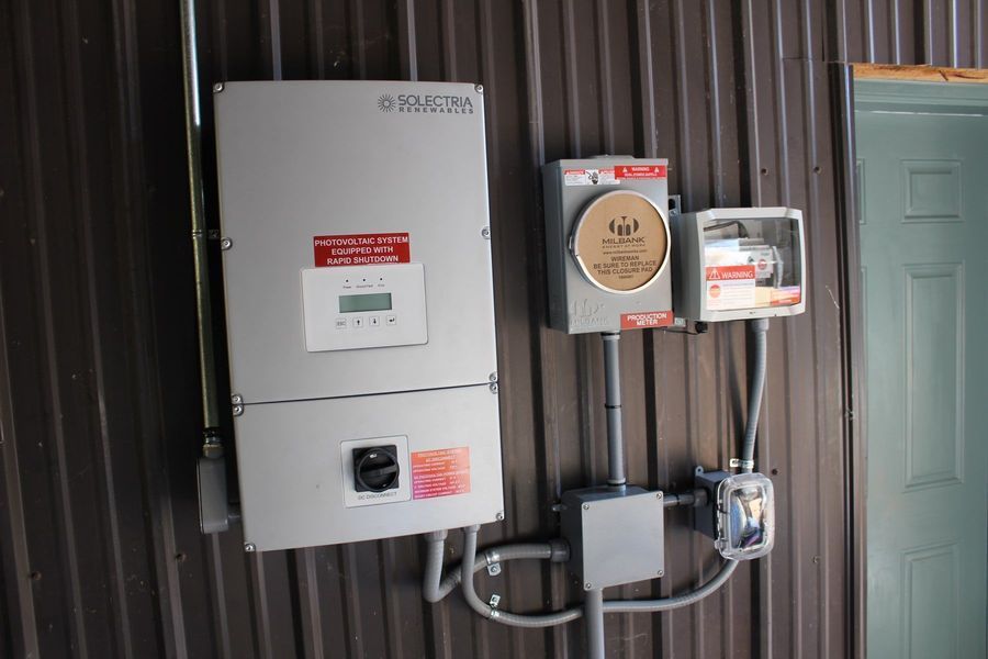 Solar power equipment mounted on a brown metal wall, including an inverter, meter, and electrical connections.