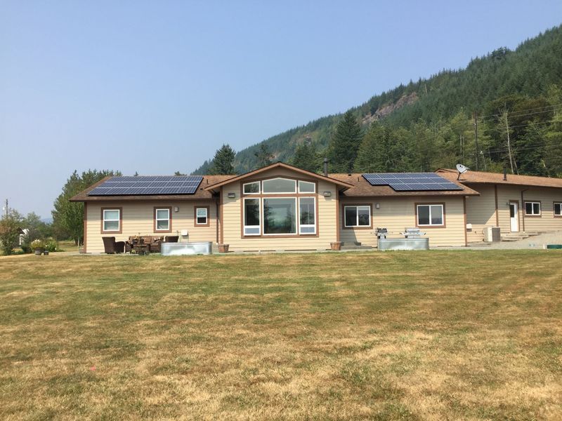 House with solar panels on roof, set on a grassy lawn with a forested hill in the background.