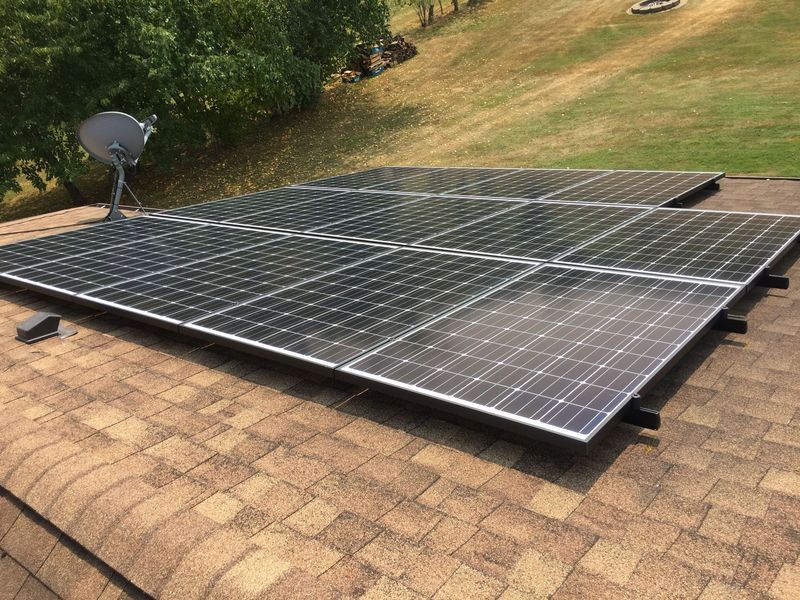 Solar panels on a shingled roof with a satellite dish and a grassy background.
