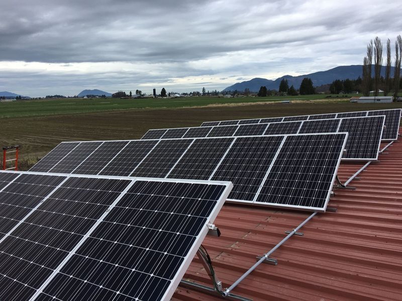 Solar panels installed on a red corrugated metal roof, with a field and mountains in the background under a cloudy sky.