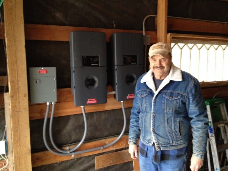Man stands near solar power equipment mounted on a wooden wall. He wears a denim jacket and a cap.