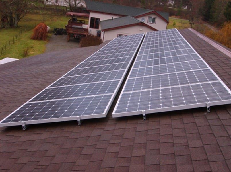 Two rows of solar panels mounted on a brown shingled roof, set against a house and landscape.