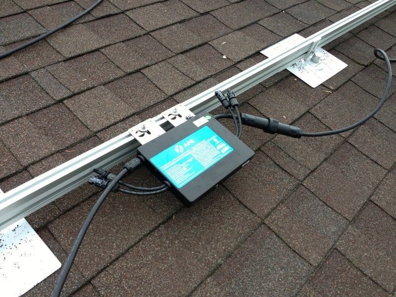 Solar panel component on a shingled roof, with black and blue box, cables, and metal supports.