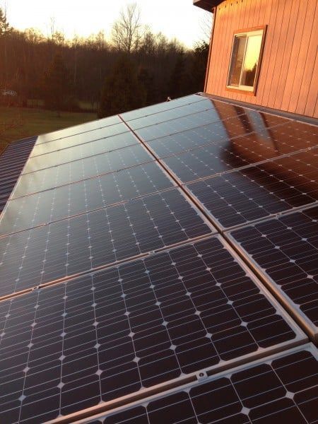 Solar panels on a sloped roof reflecting sunlight, with a house and trees in the background.