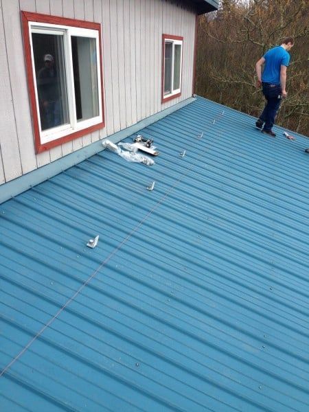 Man on a blue metal roof, near a white building with red-trimmed windows.