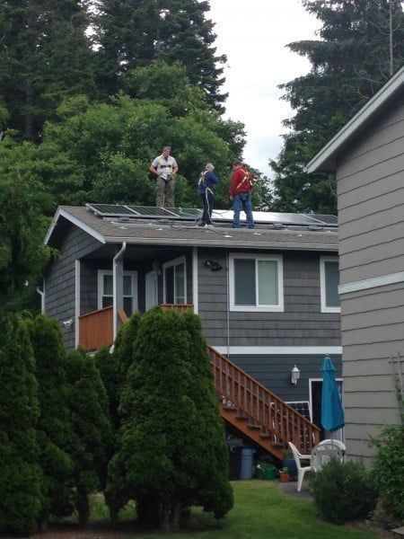 Three people on a rooftop installing solar panels. House with gray siding, lush green trees in background.