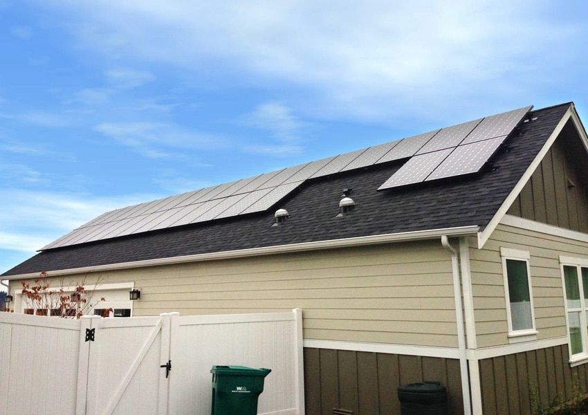 Solar panels installed on a residential roof against a blue sky. Beige siding with white fence and trash bins.