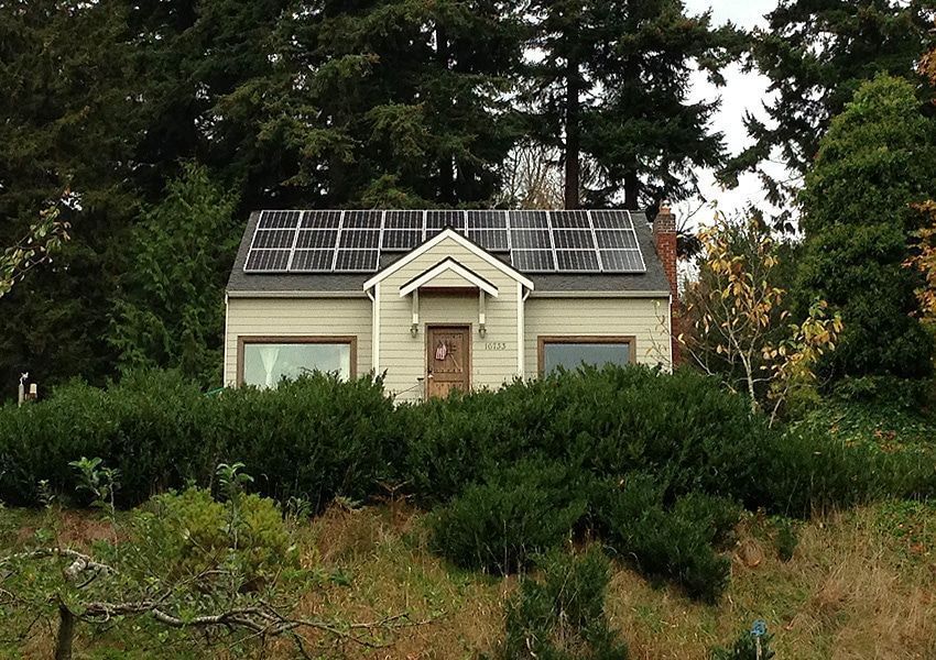 Small house with solar panels on roof, surrounded by green foliage and trees.