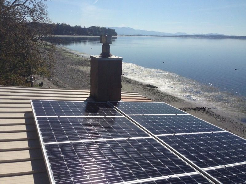 Solar panels on a roof with a chimney, overlooking a calm body of water and a beach.