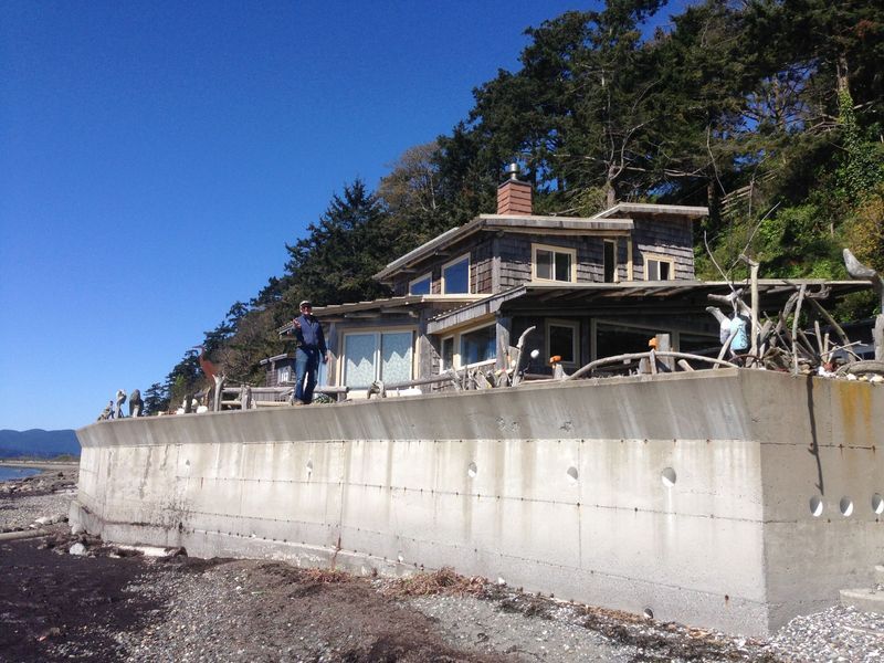 Coastal home on a concrete barrier, man stands on wall, blue sky.