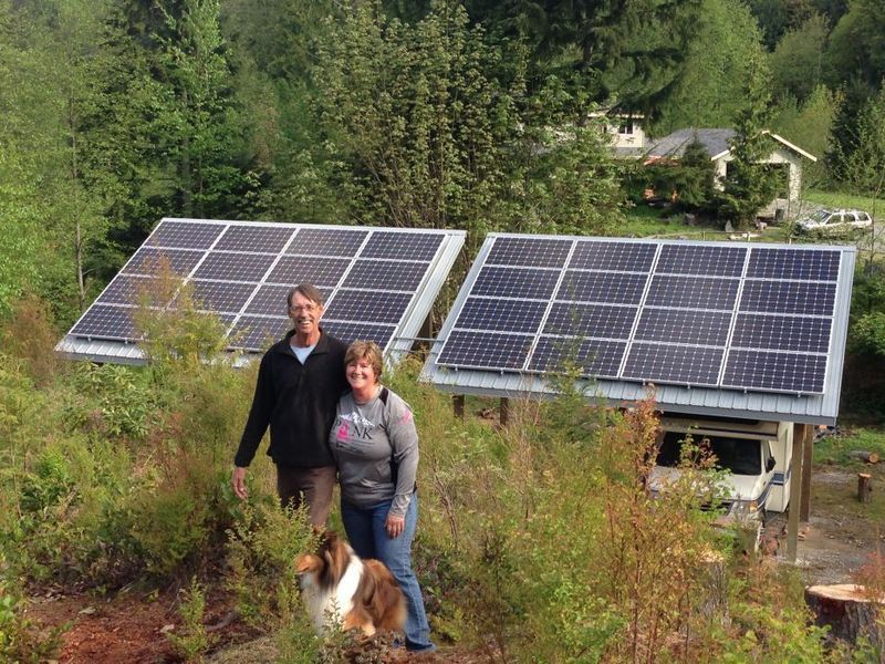 Couple and dog stand by solar panels in a wooded area. Panels sit above a vehicle.