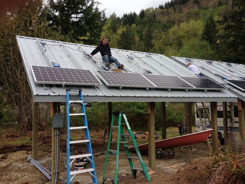 People installing solar panels on a metal roof of a wooden structure in a wooded area.