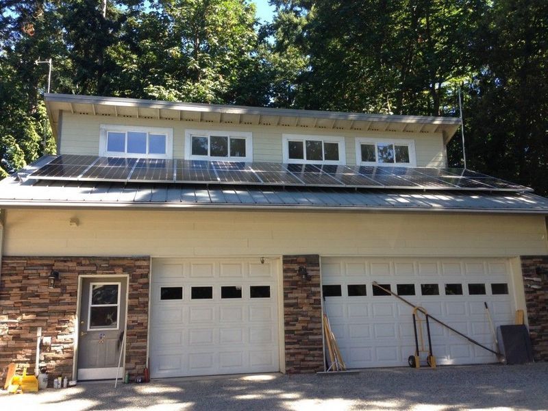 Garage with solar panels on the roof, white doors, windows above, and trees in the background on a sunny day.