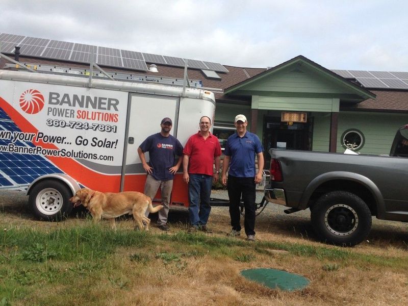 Three men stand beside a trailer and truck in front of a building with solar panels. A dog is in front.