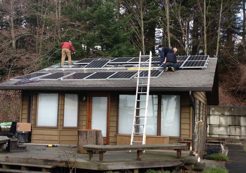 Two people install solar panels on a cabin roof; a ladder leans against the building.
