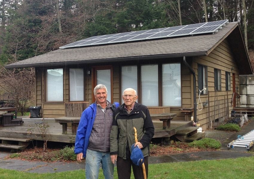 Two men stand in front of a small house with solar panels on the roof; one holds a cane and blue hat.