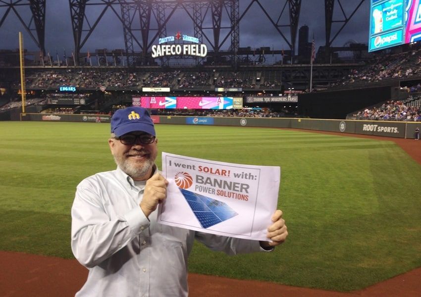 Man holding sign at Safeco Field, promoting solar energy. Stadium in background.
