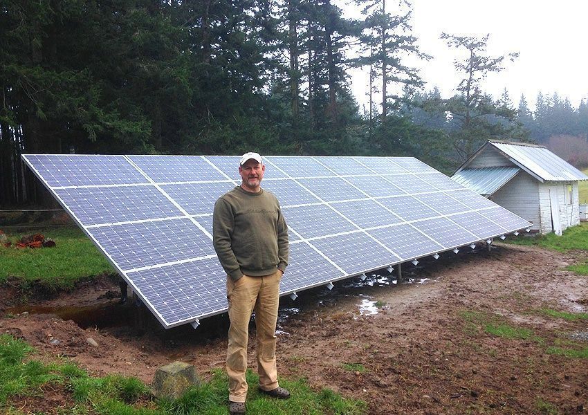 Man standing near large solar panel array on a muddy field with a small building in the background.