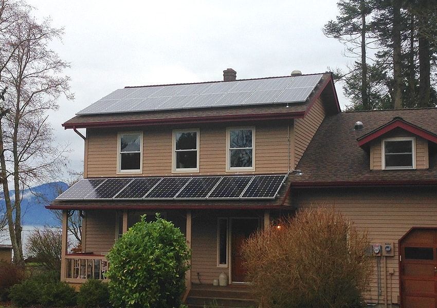 Two-story house with solar panels on roof and over porch. Tan siding, brown roof. Overcast day.