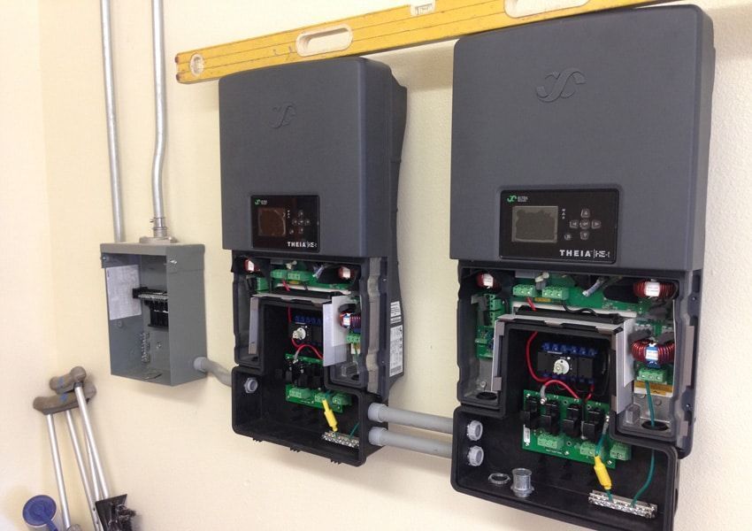 Two dark gray solar inverters mounted on a wall with electrical conduit and a circuit breaker box. A level rests above them.