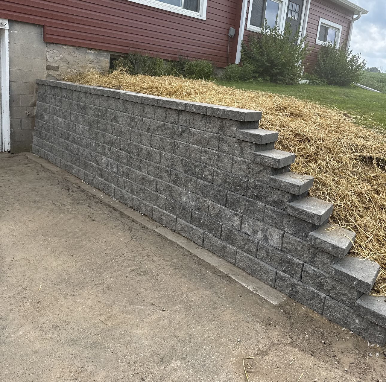 Gray retaining wall next to a cement driveway and a grassy hill with a red house in the background.