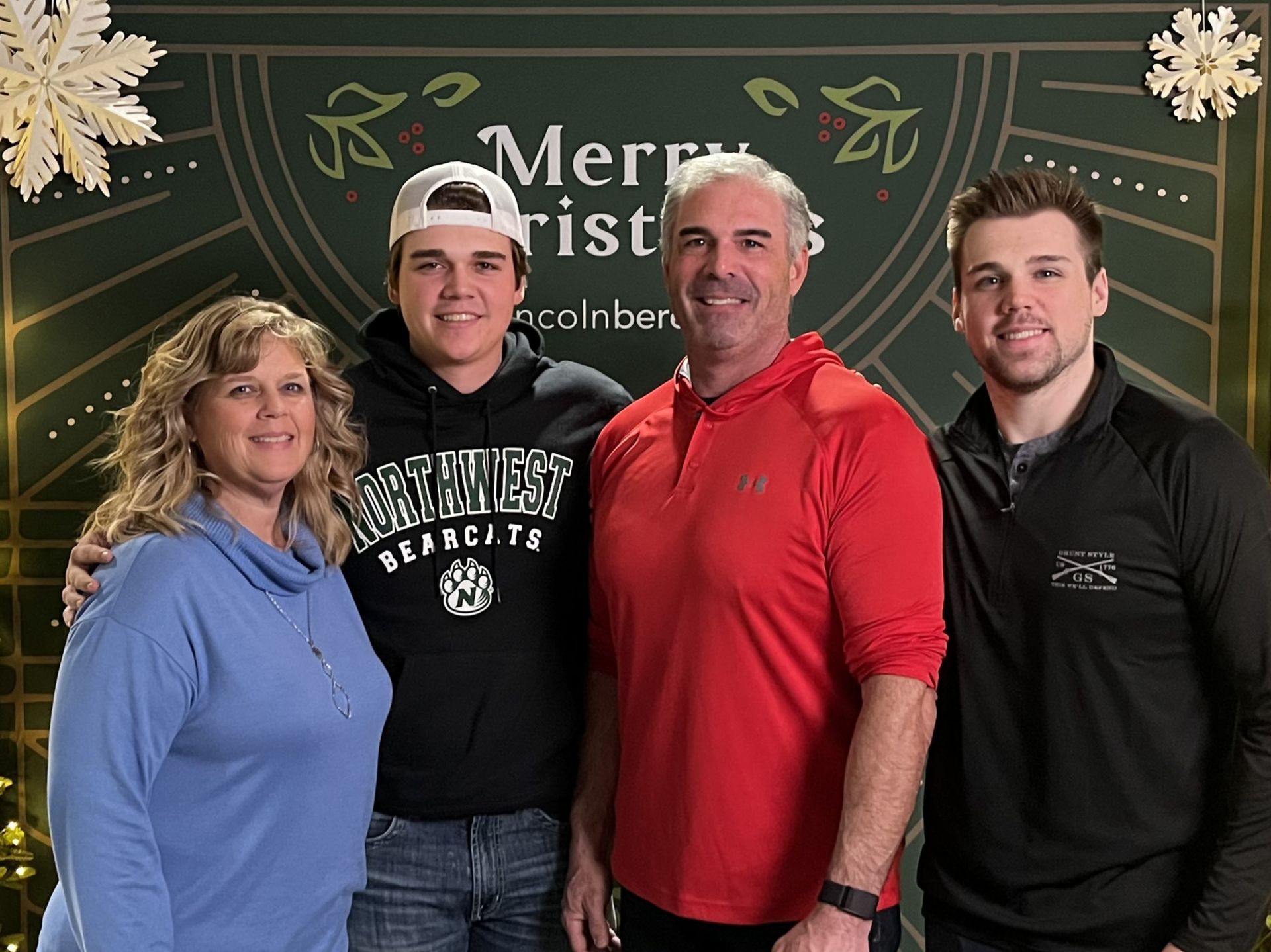Four people standing in front of a festive Merry Christmas backdrop, smiling at the camera.
