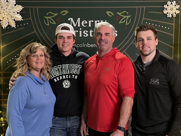 Four people standing in front of a festive Merry Christmas backdrop, smiling at the camera.