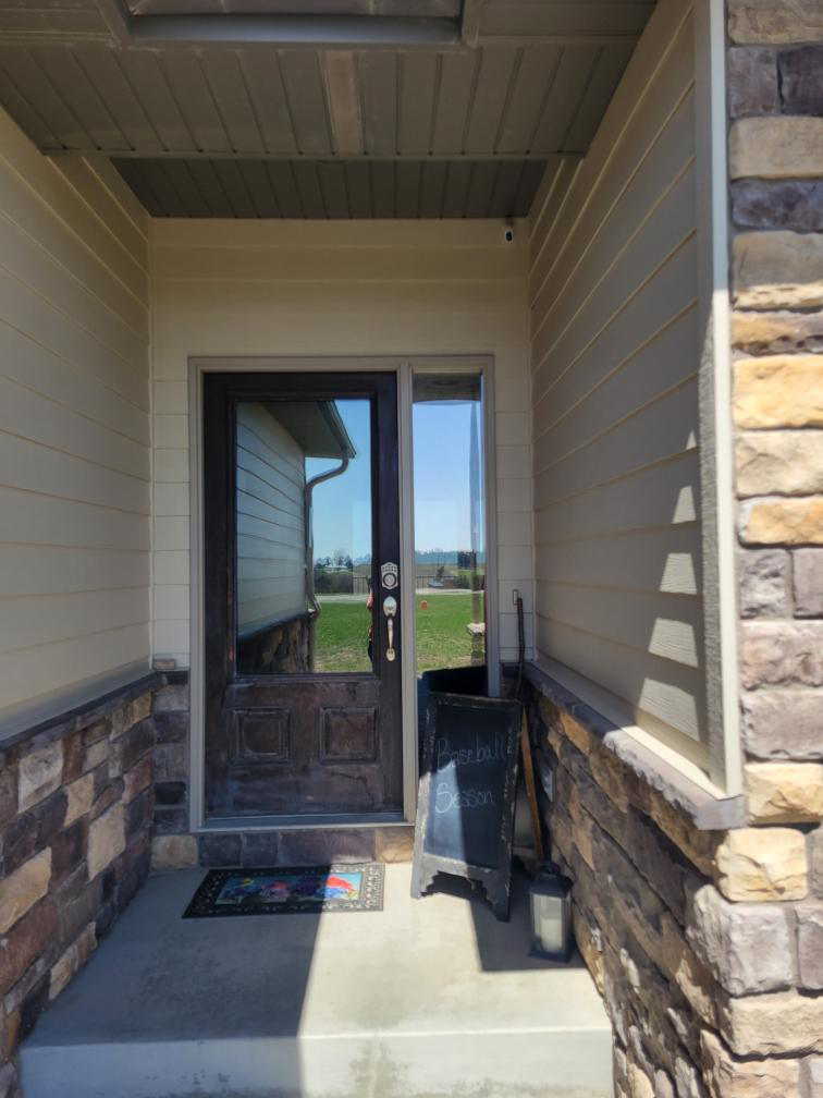 Front porch with a glass door, stone columns, beige siding, and a doormat under a covered entry.