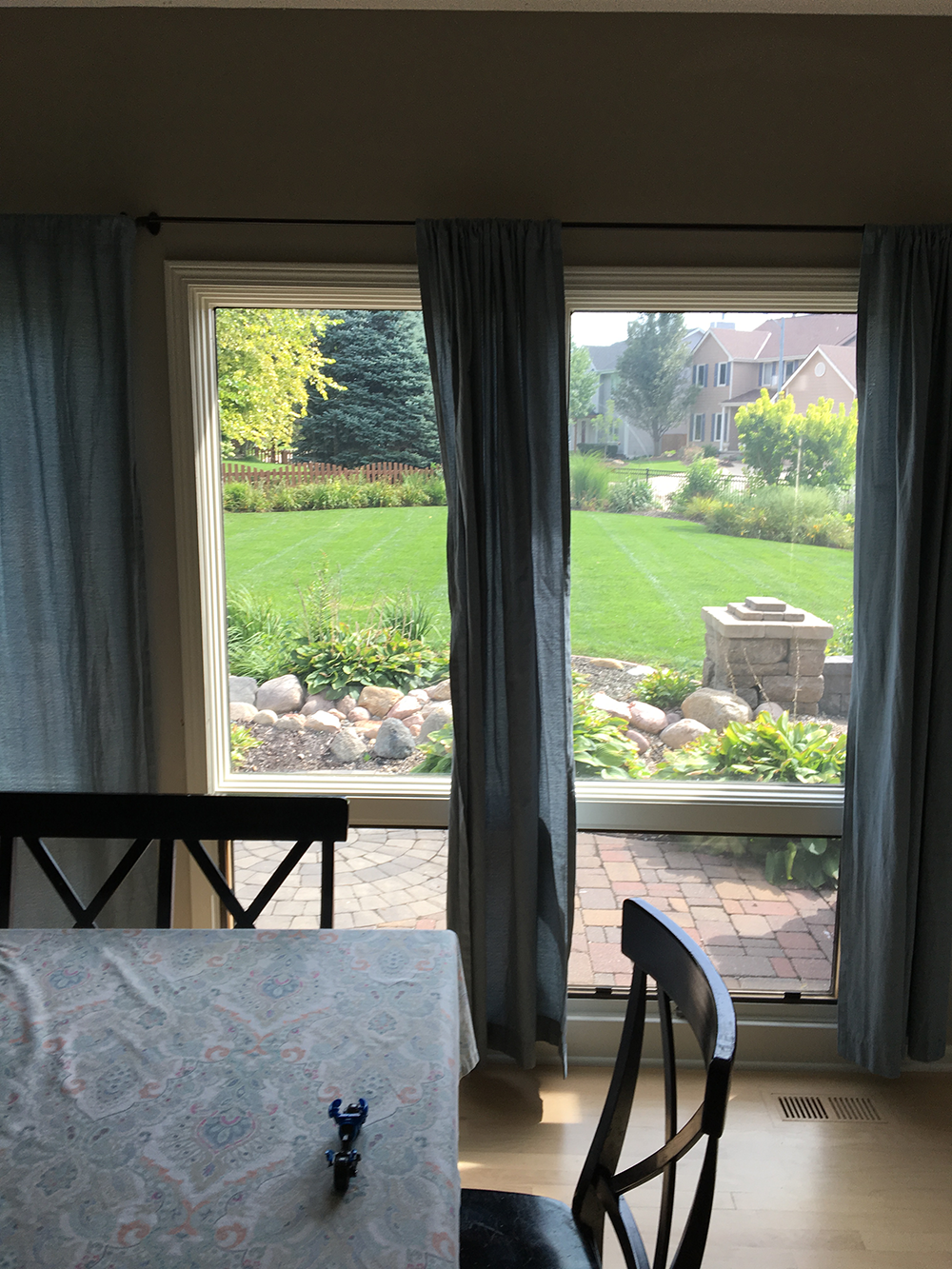 Dining area with table and chairs overlooking a green backyard through large windows and curtains