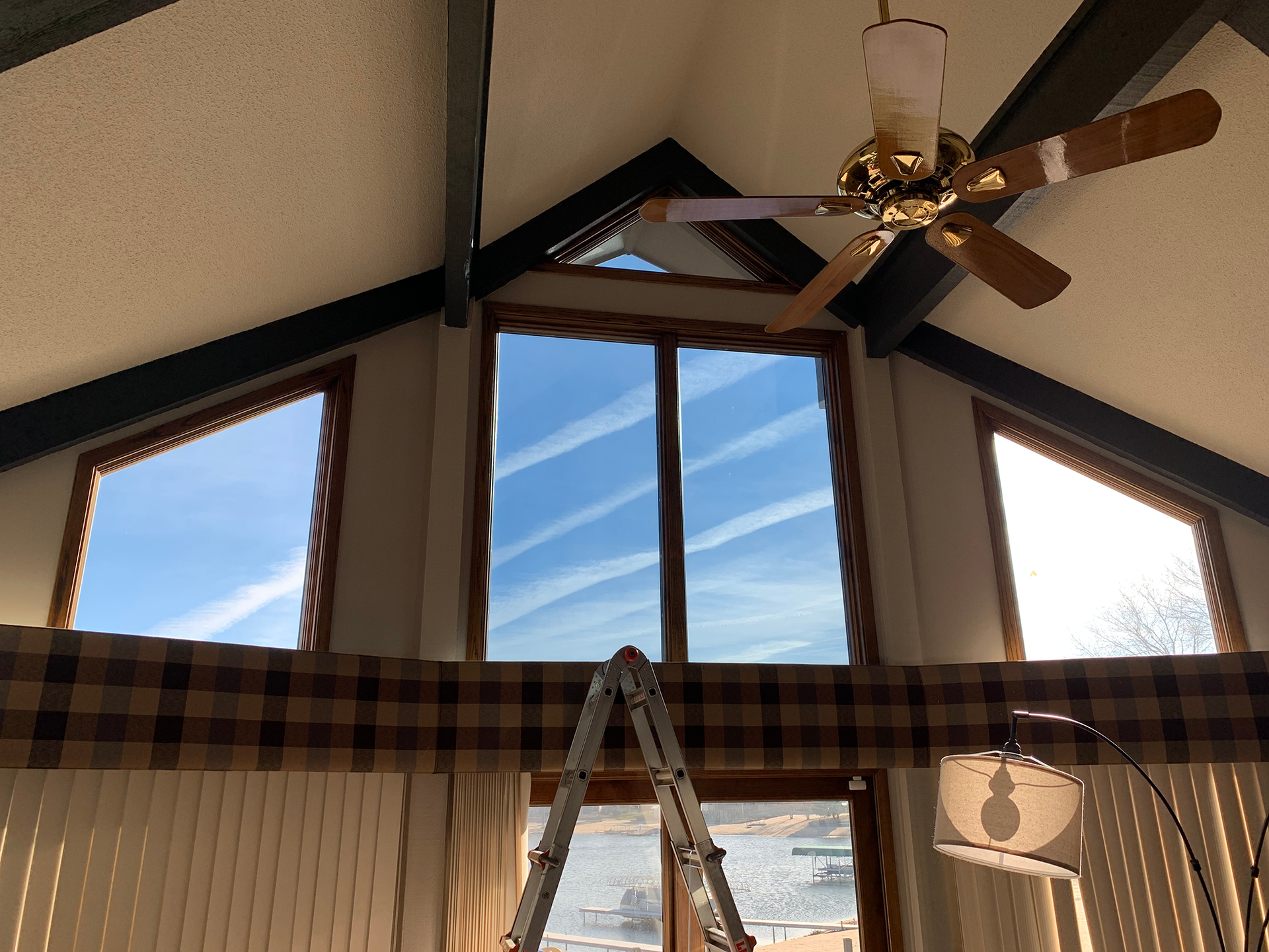 A vaulted room with a ceiling fan, three windows, a step ladder, and a plaid valance.