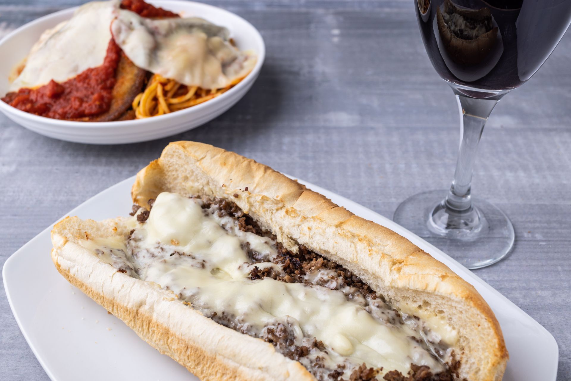 A sub sandwich with cheese and meat is on a plate next to a glass of wine.
