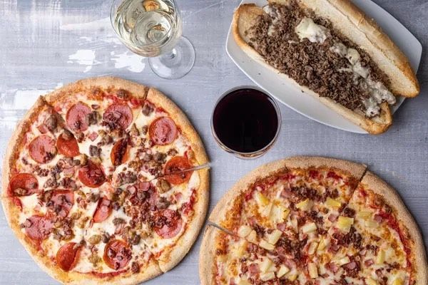 Three pizzas , a sub sandwich , and a glass of wine are on a table.