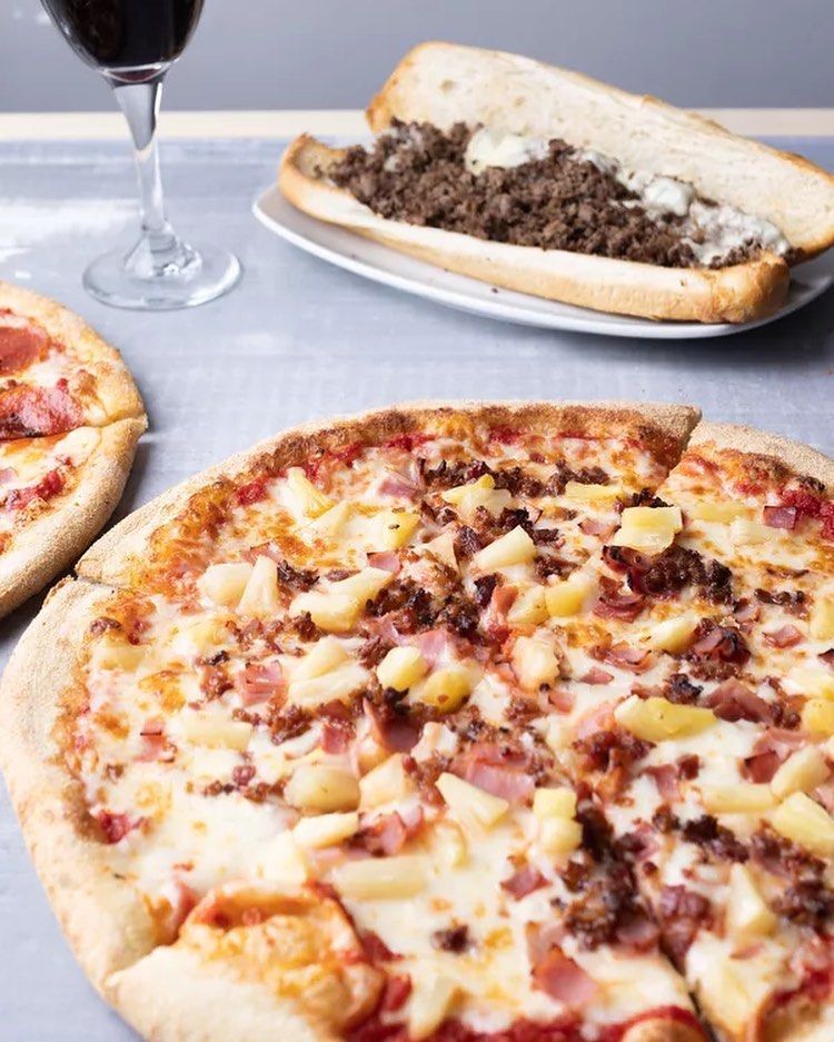 A pizza and a sub sandwich are on a table next to a glass of wine.